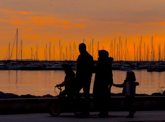 A silhouette of a family walking by the harbor at sunset with boats in the background.
