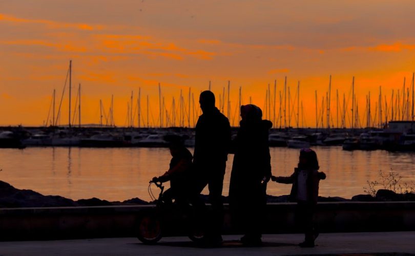 A silhouette of a family walking by the harbor at sunset with boats in the background.